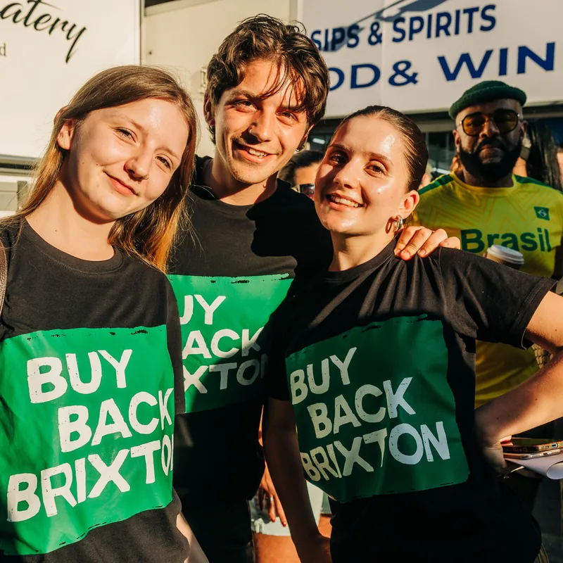 Three young supporters in Buy Back Brixton T-shirts, arms round each other, smiling at the camera.