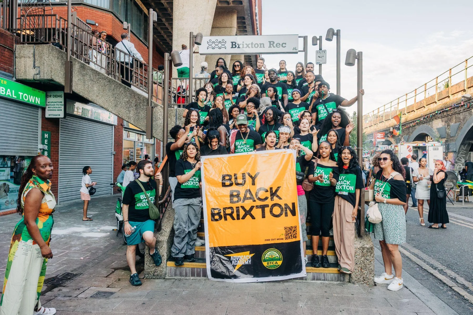 A large group of Advocacy Academy volunteers in Buy Back Brixton T-shirts on the steps at Brixton Rec, holding an orange Buy Back Brixton banner.