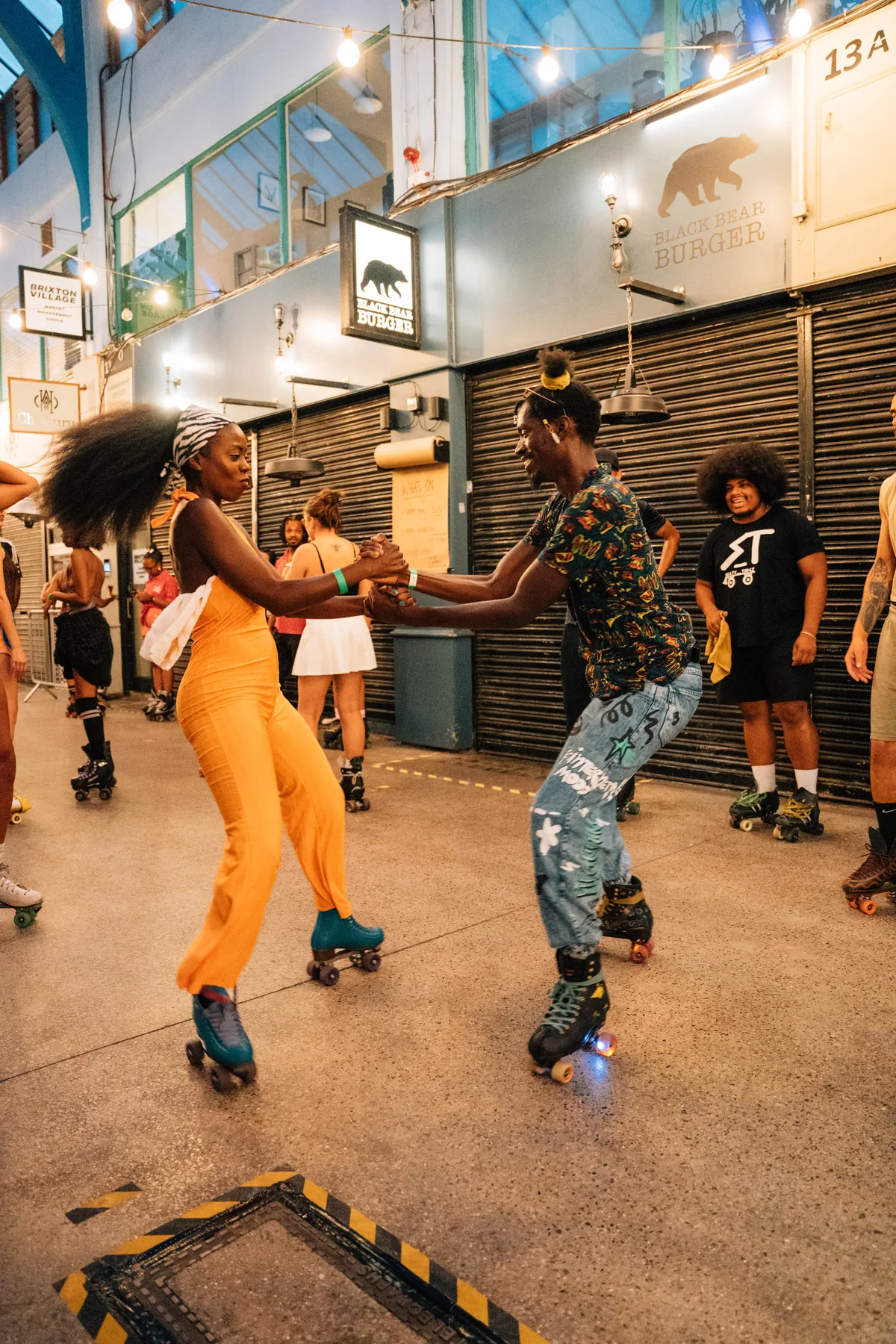 Inside Brixton Village: skaters dancing down the arcade between shuttered units and shopfronts, strings of lights overhead.