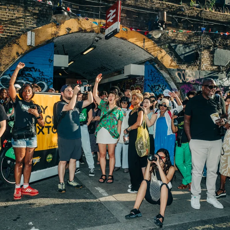 Supporters cheering with arms raised beneath the arches, an orange Buy Back Brixton banner among them.